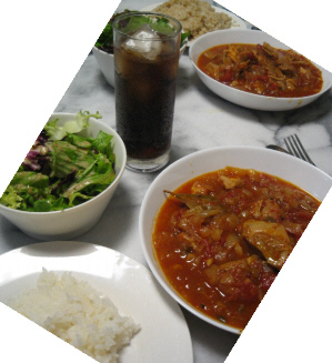 丸い白いお皿に入れられたチキンの煮込み料理。ライスとサラダも並べられています。真ん中にグラスに注がれたコーラが