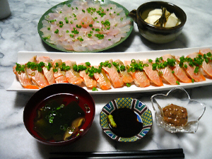 手前左から順に、味噌汁椀、お醤油の小皿、花形の透明な小鉢に茶色いものが入っています。真ん中の細長いお皿には、サーモンの握り寿司が、青ネギを上に散らしてあります。一番向こう側には、白身のお刺身が、お皿一杯に並べられています。その隣には、大根の漬物が入った小鉢が。
