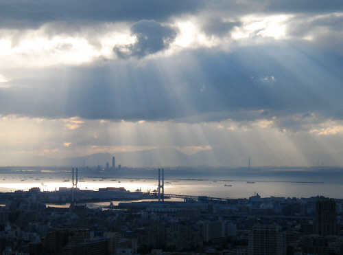 全体がグレーと沈んだブルーの風景。街並みの向こうに部分的に白く光る海が広がって、厚い雲の切れ間から太陽の光がうっすらと射しています。光のカーテンがオーロラのように出来て、色合いの無い世界が一層幻想的に見えています。