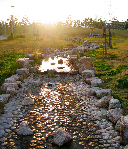 公園内に作られた人工の川の水辺。水が殆ど無く、少し残った水溜りに夕日が反射してまばゆく光っています。細い人工の川がまるで小道のように見えて、ずっと続く先は夕日の中に消えているように見えます。