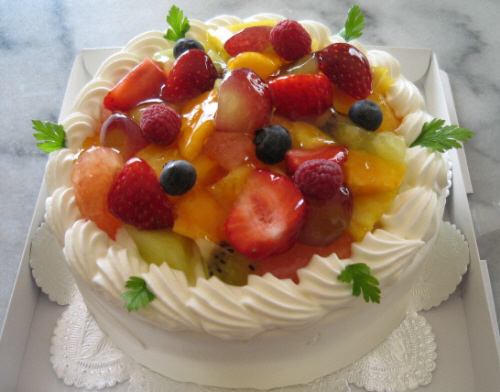 上に色々なフルーツが乗ったホールのケーキです。生クリームの白がフルーツの赤い色合いを引き立てています。周りに飾られたチャービルの緑が綺麗です。