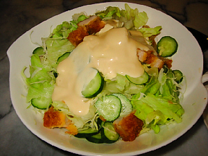 白い大きめの深い更に入ったサラダ。ところどころに茶色い角切りのものが見えているのは、とんかつを切ったもののようです。マヨネーズのようなドレッシングがかかっています。