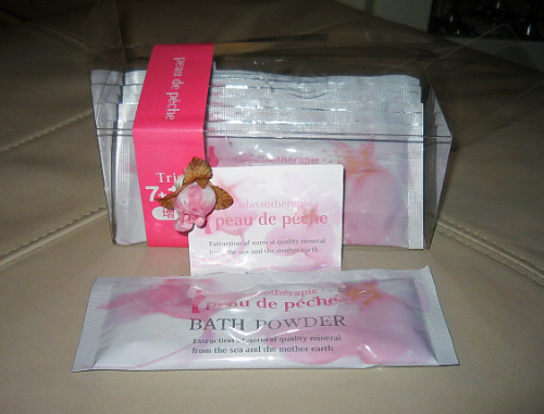 透明のケースに入った入浴剤。ピンクの（たぶん）桃の花が描かれており、全体にピンクがかっています。袋状のパッケージが7個入ったもの。peau de Peche BATH POWDER　と書かれてあります。