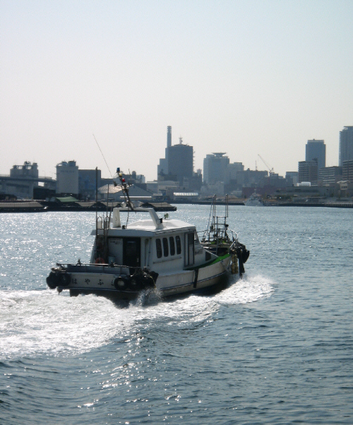 川のように見える入り江、でもここは海です。釣船が白い波を立てて走っています。船の向こう側に神戸市内の高いビル軍が見えています。