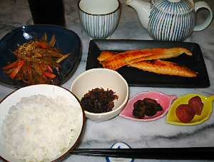 ご飯、ブルーの器にきんぴらごぼう、小さな白い丸の器に、昆布の振りかけ、葉っぱの形の小さな器に、梅干ときゅうりのきゅうちゃんがそれぞれ、長方形の黒いお皿にはらす、その向こうにはおそろいの縞模様の急須と湯飲み茶碗が。