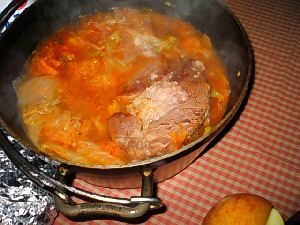 完成し、ダッチオーブンの蓋を取ったスープのアップ画像。ギンガムチェックのテーブルクロスの上にお鍋が置かれています。色が赤いのはキムチのせいですね。大きな肉の塊が見えています。
