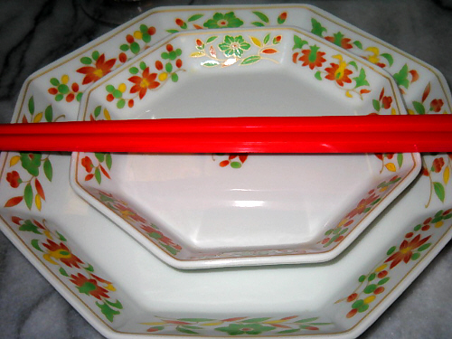 取り分けざらのアップ画像。八角形の大小のお皿、白いお皿の淵に赤と緑の小花模様が描かれ、金色の縁取りがしてあります。大小二枚重ねて、更にその上に真っ赤なやや大きめのプラスチックのお箸を乗せてあります。