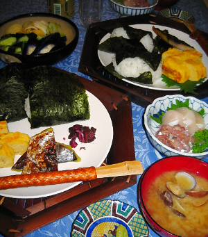 六角形の竹で作られた器の上に白い丸いお皿が乗せられ、その上におにぎり二個、焼き魚、玉子焼き、柴漬けが盛られています。竹の箸は竹で編んだ筒状の箸袋に収められて、お皿の上に渡してあります。アサリの味噌汁椀、刺身の盛られた小さめの丸い器、そして、茄子、きゅうり、白菜の漬物が盛り込まれた大きめの器が奥に見えています。