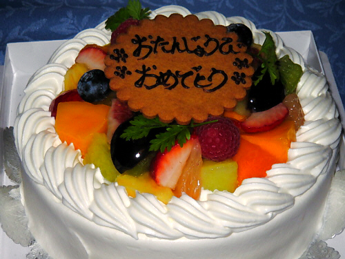 真っ白な生クリームで包まれたホールのケーキ。色とりどりのフルーツがたっぷり乗っていて、その周りはクリームのデコレーションで囲まれています。真ん中におたんじょうびおめでとうと書かれたクッキープレートがどんと乗っかっています。