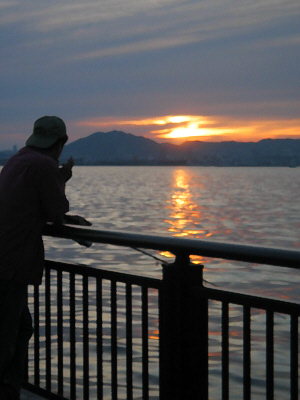 釣りざをを垂らしている男性のシルエットの向こうに、山間に沈みかけている夕日が海に反射して、オレンジのラインを作っています。穏やかな神戸港の夕暮れの一こま。