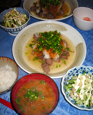 ご飯、味噌汁、小鉢に入ったきゅうりのサラダ、それらの向こうに大皿に盛られたサイコロステーキ、たっぷり大根おろしとわけぎが乗っています。