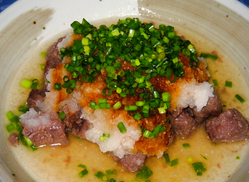丸い和風の大き目の更に盛られたサイコロステーキのアップ画像。角切りのお肉の上に大根おろしがどっさり、そして緑のネギ。ポン酢がかかって大根おろしにお醤油色のグラデーションが。